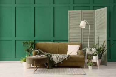 Oturma odasının içinde rahat bir kanepe, lamba, puf ve çiçekleri var.