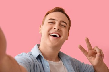Genç adam zafer jesti yapıyor ve pembe arka planda selfie çekiyor, yakın plan.