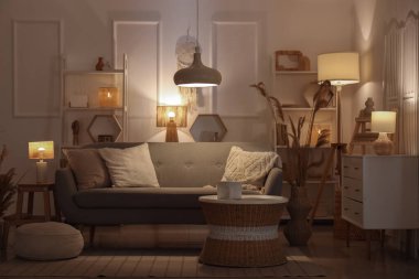 Rahat bir kanepe, çekmeceler, kahve masası, otlar ve oturma odasının içinde parlayan lambalar.