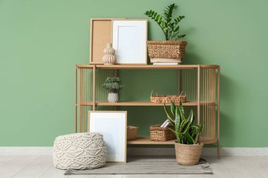 Raflar, ev bitkileri, pouf ve çerçeveler yeşil duvarın yanında.