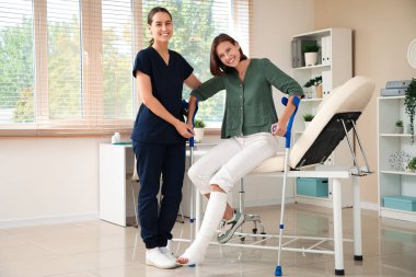 Kadın doktor, kırık bacaklı ve koltuk değnekleriyle klinikteki koltuktan kalkmasına yardım ediyor.