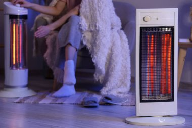Akşam evde, battaniyeli genç bir kadın elektrikli ısıtıcıyı açıyor.