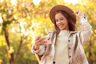 Cep telefonlu şirin kız, sonbahar günü parkta selfie çekiyor.