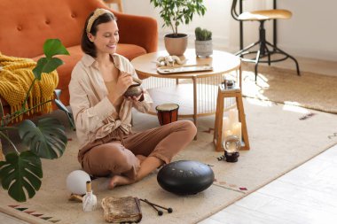 Young woman with Tibetan singing bowl and glucophone meditating on carpet at home clipart