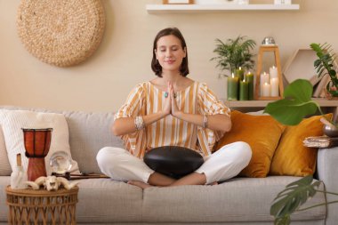 Glukofon kullanan genç bir kadın evdeki gri kanepede meditasyon yapıyor.