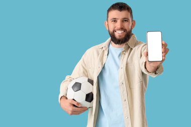 Mavi arka planda futbol topu ve cep telefonu olan yakışıklı genç bir adam. Spor bahsi kavramı