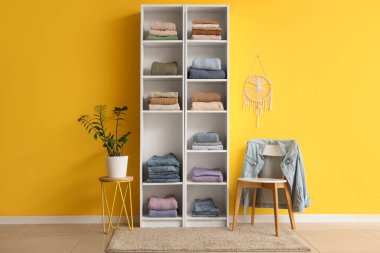 Katlanmış elbiseli raf ünitesi, sandalye ve çiçek renkli duvarın yanında.