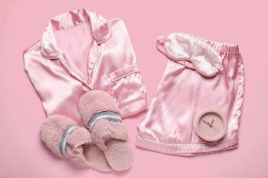 Uyku maskeli ipek pijamalar, pembe arka planda terlikler ve çalar saat.