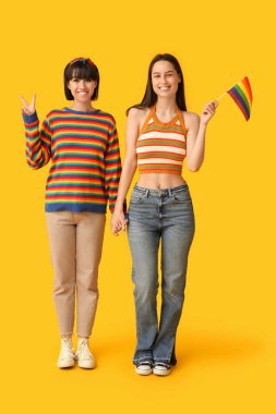 LGBT bayrağı taşıyan genç lezbiyen çift sarı arka planda zafer jesti gösteriyor