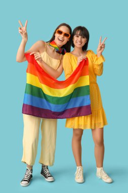 LGBT bayrağıyla genç lezbiyen çift mavi arka planda zafer jesti gösteriyor