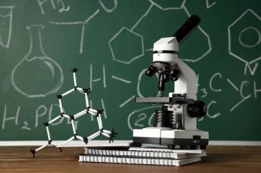 Modern mikroskop, moleküler model ve kimyasal formüllü karatahtanın yanındaki ahşap masadaki kopyalar.