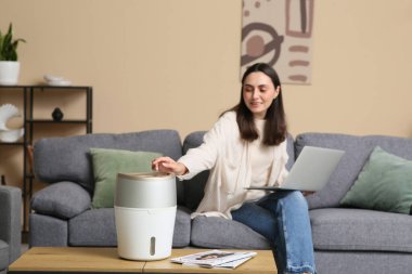 Dizüstü bilgisayarı ve modern hava nemlendiricisi olan güzel bir kadın evdeki kanepede oturuyor.