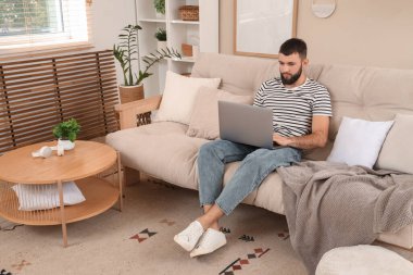 Genç sakallı adam evdeki kanepede dizüstü bilgisayarla çalışıyor.