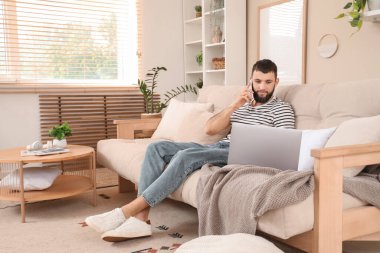 Bilgisayarlı genç sakallı adam evdeki koltukta cep telefonuyla konuşuyor.