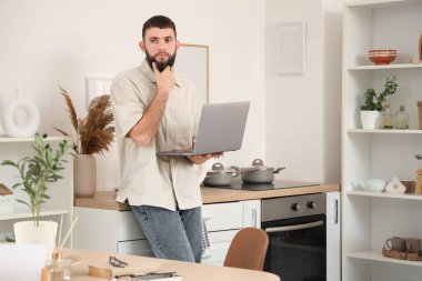 Mutfakta dizüstü bilgisayarla çalışan düşünceli genç sakallı adam.