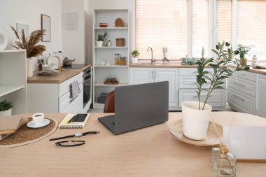 Mutfaktaki yemek masasında cep telefonu, kol saati ve kahve fincanı olan dizüstü bilgisayar.