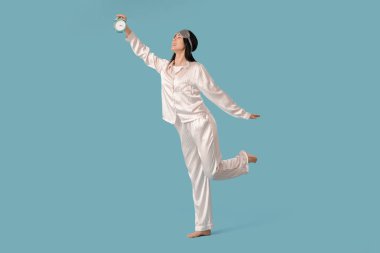 Mavi arka planda uyku maskesi ve çalar saati olan güzel genç bir kadın.