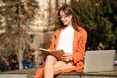 Not defteri ve dizüstü bilgisayarı olan genç bir kadın şehirde bankta oturuyor.