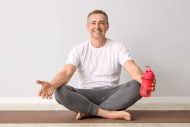 Elinde su şişesi olan olgun bir adam yoga minderinde, ışık duvarının yanında oturuyor.