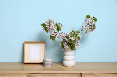 Mavi duvarın yanındaki masada çiçek açan dalları olan bir vazo.