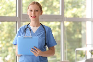 Kliniğin penceresinin yanında steteskop ve panosu olan mutlu bir kadın doktor.