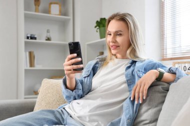 Genç bir kadın kanepede oturuyor ve oturma odasında cep telefonu kullanıyor.