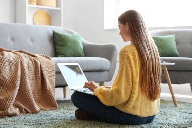 Genç bir kadın dizüstü bilgisayar kullanıyor ve oturma odasında yerde oturuyor.