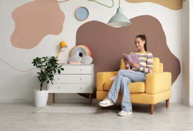 Çocuk odasındaki koltukta oturmuş kitap okuyan genç bir kadın.