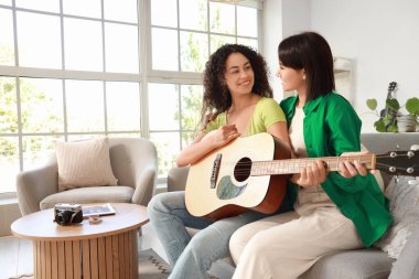 Genç lezbiyen çift evdeki kanepede gitar çalıyor.