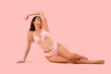 Güzel, genç, mutlu, Afro-Amerikalı, zarif dantel iç çamaşırlı, pembe arka planda oturan kadın.