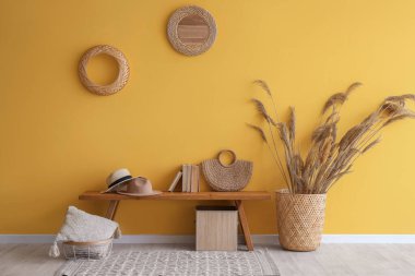 Kitaplı ahşap bank, otlar ve sarı duvarın yanında hasır dekor.