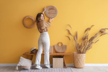 Koridordaki sarı duvara ayna asan genç kadın.