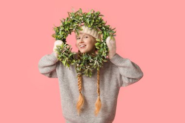 Pembe arka planda Noel çelengi olan sıcak kış kıyafetleri içinde güzel, genç ve mutlu bir kadın.