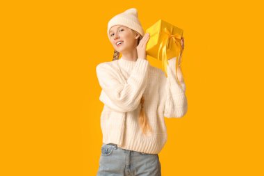 Sarı arka planda hediye kutusuyla kalın kış kıyafetleri içinde güzel bir kadın.