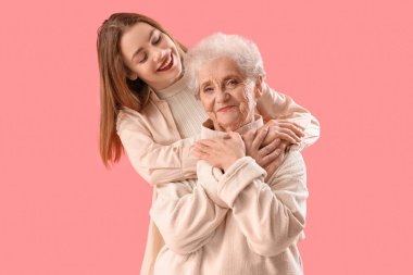 Genç bir kadın pembe arka planda büyükannesine sarılıyor.