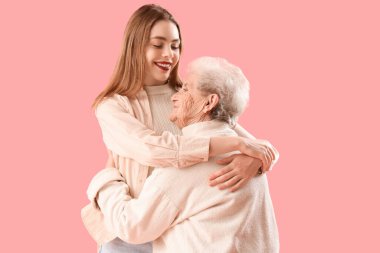 Genç bir kadın pembe arka planda büyükannesine sarılıyor.