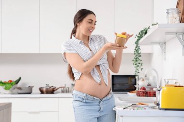 Genç hamile kadın mutfakta fıstık ezmesi kavanozu açıyor.