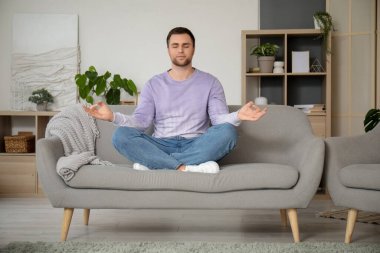 Genç adam evdeki gri kanepede meditasyon yapıyor.