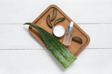 Aloe vera yaprağı, ahşap tepsi, kavanoz ve beyaz ahşap arka planda bir şişe kozmetik ürünü.