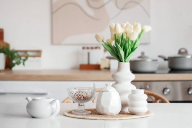 Mutfağın içinde çaydanlık ve vazo çiçekli beyaz yemek masası.