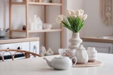 Mutfağın içinde çaydanlık ve vazo çiçekli beyaz yemek masası.