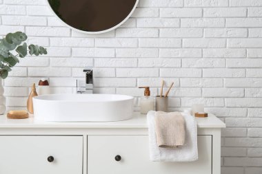 Modern lavabo, banyo malzemeleri ve banyoda beyaz tuğla duvarın yanında yeşil dallı vazo.