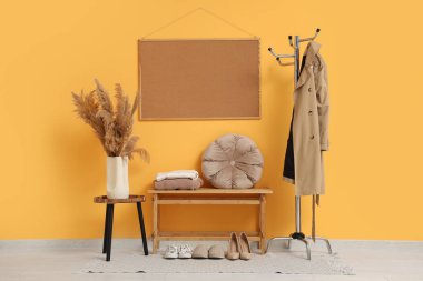 Yedek kulübesi, trençkotlu askı vazoda otlar ve sarı duvardaki çerçeve.