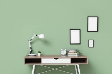 Ofiste yeşil duvarın yanında saati, lambası ve kitapları olan şık bir masa.