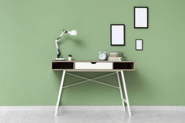 Ofiste yeşil duvarın yanında saati, lambası ve kitapları olan şık bir masa.