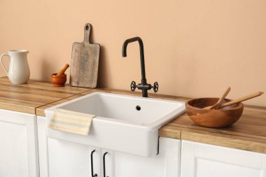 Mutfakta bej duvarın yanındaki ahşap tezgahta seramik lavabo.