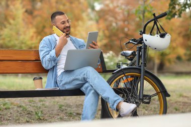 Bilgisayarlı, tabletli ve bisikletli genç mutlu adam parkta bankta cep telefonuyla konuşuyor.
