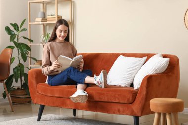 Oturma odasında kanepede oturmuş kitap okuyan genç bir kadın.