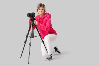 Gri arka planda oturan profesyonel kameralı güzel, genç, mutlu bayan fotoğrafçı.