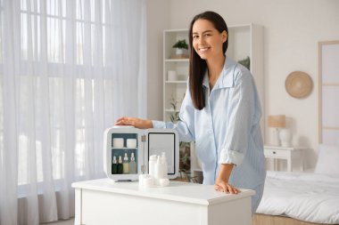 Evinde küçük kozmetik buzdolabı olan, masanın yanındaki mutlu genç kadın.